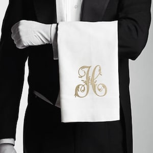 May include: A white linen towel with a gold embroidered initial "H" is held by a person in formal attire. The person is wearing a black tuxedo, white gloves, and a white bow tie. The towel is the focal point.