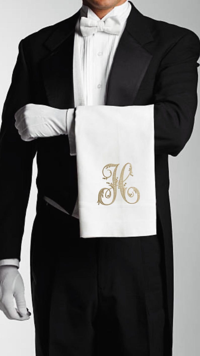 May include: A white linen towel with a gold embroidered initial "H" is held by a person in formal attire. The person is wearing a black tuxedo, white gloves, and a white bow tie. The towel is the focal point.