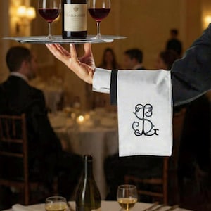 May include: A waiter holds a silver tray with a bottle of red wine and two wine glasses filled with red wine. A white napkin with black embroidery is draped over the waiter's arm. The scene is set in a restaurant.