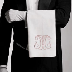 May include: A formal setting with a person in a tuxedo holding a white towel. The towel features an ornate, light-red monogram. The person is wearing white gloves and a bow tie, suggesting a service or hospitality theme.
