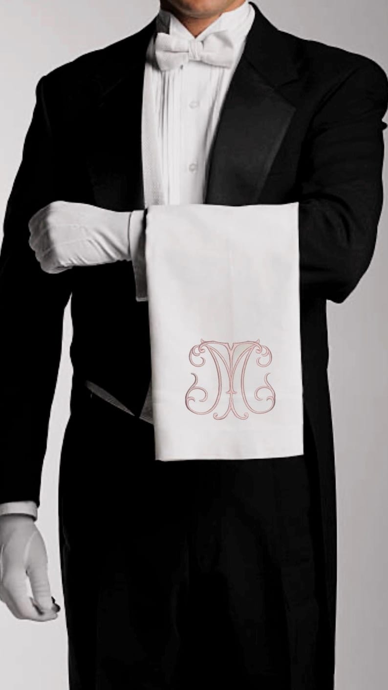 May include: A formal setting with a person in a tuxedo holding a white towel. The towel features an ornate, light-red monogram. The person is wearing white gloves and a bow tie, suggesting a service or hospitality theme.