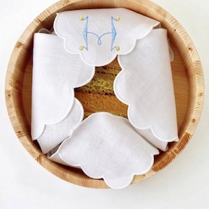 Puede incluir: Un cuenco redondo de madera que contiene una servilleta de lino blanco con bordes festoneados. La servilleta está bordada con una letra H azul y pequeños puntos amarillos, doblada para revelar parcialmente un alimento texturizado marrón claro.