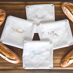 May include: Four white linen napkins with scalloped edges, each embroidered with a different word: "Arepas", "Pan", "Casabe", and "Casabe". The napkins are arranged on a wooden surface with loaves of bread.