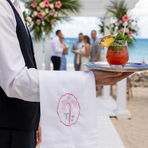 May include: A waiter holds a silver tray with a cocktail garnished with pineapple and mint. A white linen towel with a pink palm tree and initials is also held. The background shows a beach wedding with floral decorations.