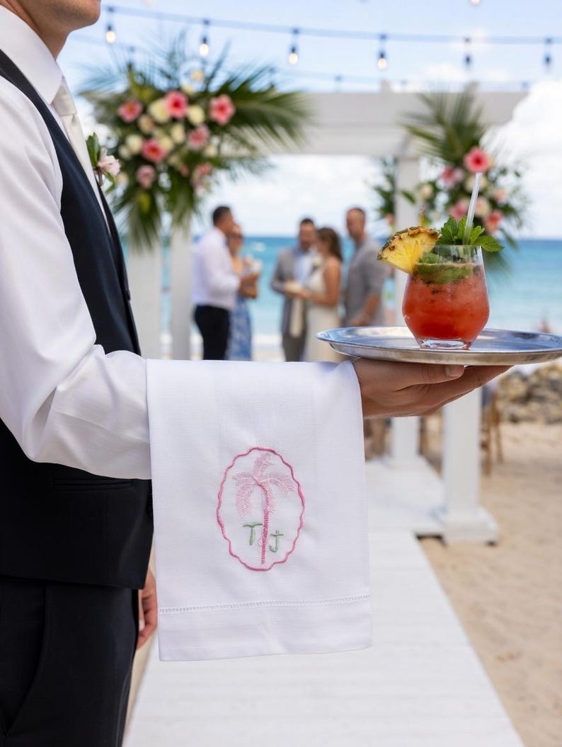 May include: A waiter holds a silver tray with a cocktail garnished with pineapple and mint. A white linen towel with a pink palm tree and initials is also held. The background shows a beach wedding with floral decorations.