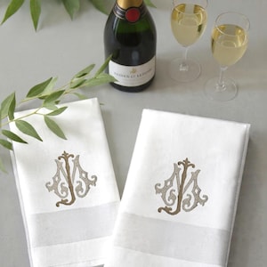 May include: Two white monogrammed napkins with a champagne bottle and two glasses of champagne. The napkins have a decorative border and a brown and tan monogram. Green leaves are in the background.