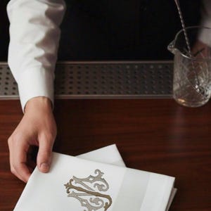 May include: White linen napkins with a decorative monogram in shades of brown and gray. The napkins have a striped border and are neatly folded. The image suggests a bar or restaurant setting.