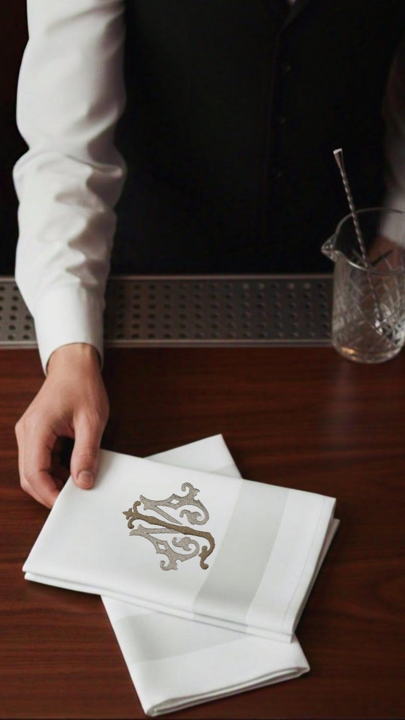 May include: White linen napkins with a decorative monogram in shades of brown and gray. The napkins have a striped border and are neatly folded. The image suggests a bar or restaurant setting.
