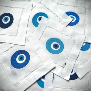 May include: A stack of white linen napkins with blue and white evil eye designs embroidered on them.