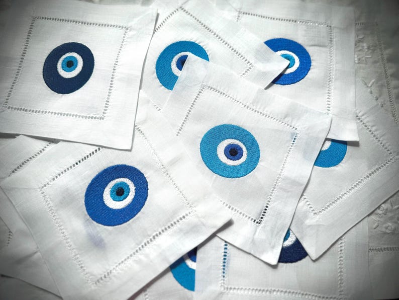 May include: A stack of white linen napkins with blue and white evil eye designs embroidered on them.