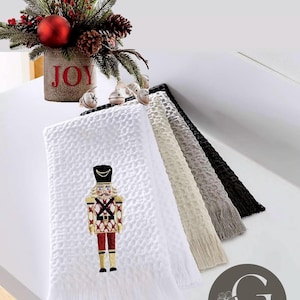 May include: Set of five white waffle weave kitchen towels with fringed edges. The towels feature a festive embroidered nutcracker design. The towels are stacked with the top towel showing the nutcracker design. The towels are labeled "Accents by Geisha".