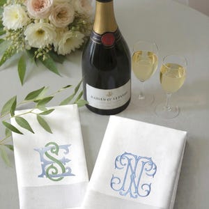 May include: A still-life image featuring a champagne bottle, two filled glasses, and two white monogrammed hand towels. A bouquet of flowers sits to the left. The towels have embroidered initials in blue and green.