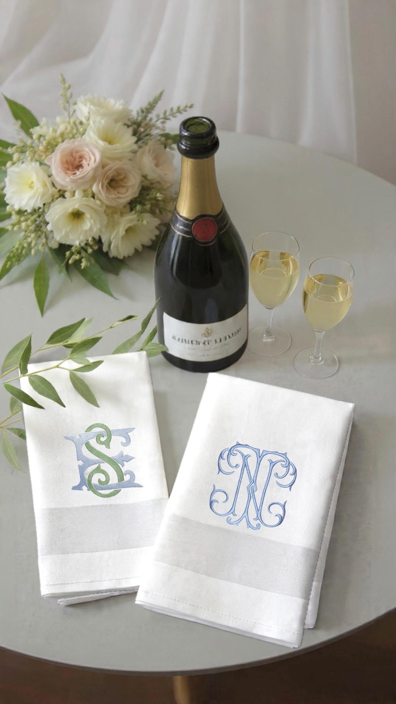 May include: A still-life image featuring a champagne bottle, two filled glasses, and two white monogrammed hand towels. A bouquet of flowers sits to the left. The towels have embroidered initials in blue and green.