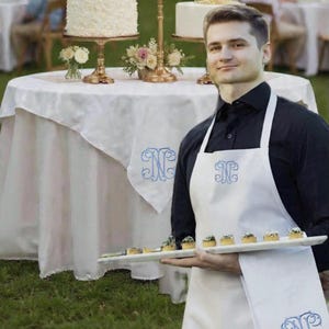 May include: A server in a white apron and black shirt holds a tray of appetizers. Three tiered cakes with floral decorations sit on a table with a white tablecloth. The apron and tablecloth have a blue monogram.