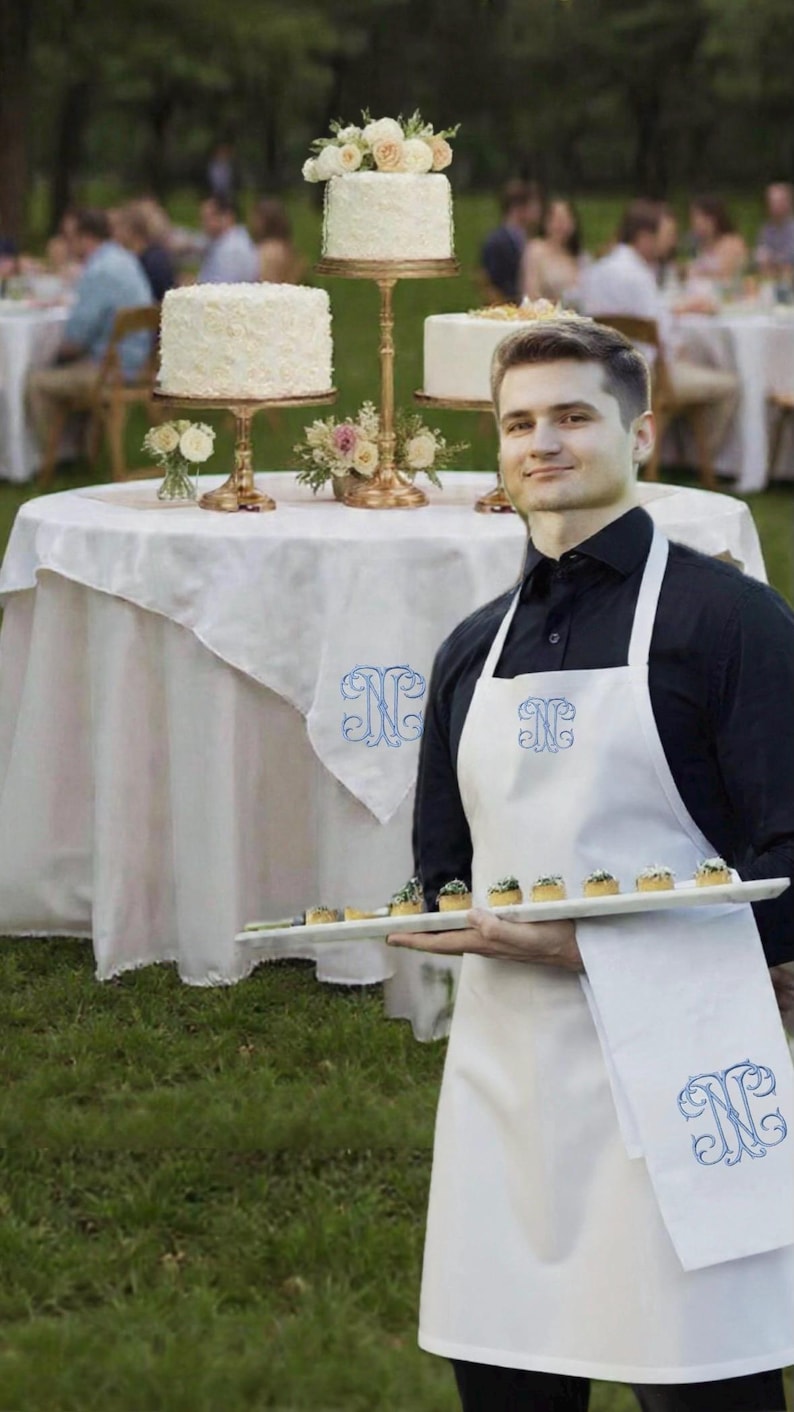 May include: A server in a white apron and black shirt holds a tray of appetizers. Three tiered cakes with floral decorations sit on a table with a white tablecloth. The apron and tablecloth have a blue monogram.