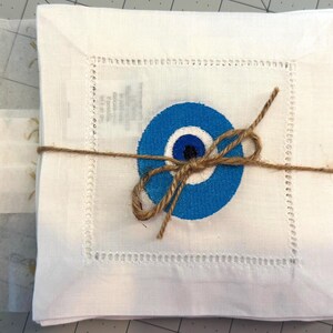 May include: A set of white linen napkins with a blue evil eye design embroidered on each napkin. The napkins are tied together with twine.