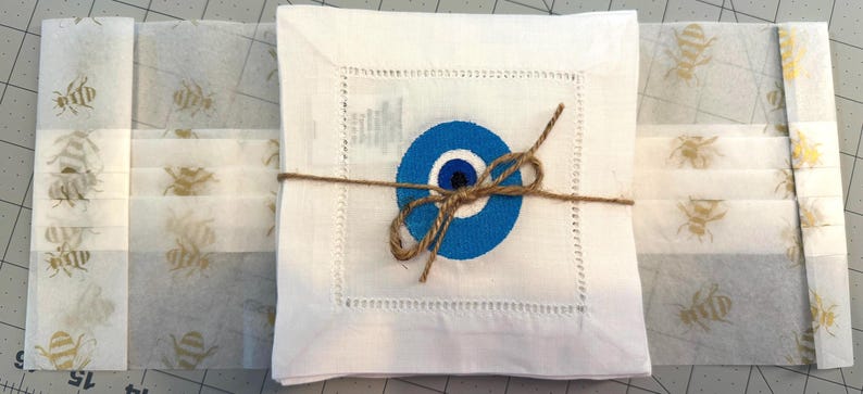 May include: A set of white linen napkins with a blue evil eye design embroidered on each napkin. The napkins are tied together with twine.