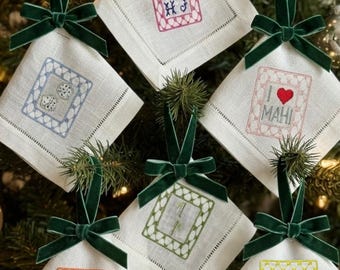 May include: Six square white linen ornaments with hemstitched edges, each featuring a dark green velvet ribbon bow. Embroidered designs include a pink frame, blue dice, a red heart with text I LOVE MAHI, a green monogram, an orange flower, and a purple dragon.