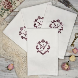 May include: White cotton towels with embroidered burgundy initials within an ornate frame. The towels are folded and arranged on a light wood surface, with decorative elements like a vintage key and dried flowers.
