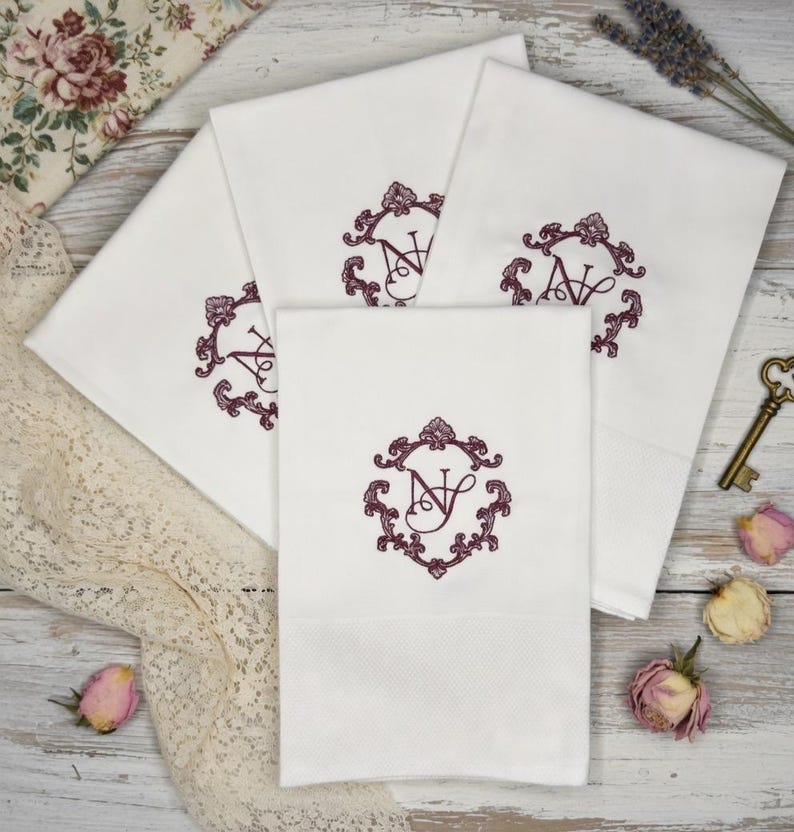 May include: White cotton towels with embroidered burgundy initials within an ornate frame. The towels are folded and arranged on a light wood surface, with decorative elements like a vintage key and dried flowers.