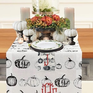 May include: A fall-themed table setting featuring a white table runner with black pumpkin illustrations. The centerpiece includes a floral arrangement, candles, and decorative pumpkins. A plate with a green border is also visible.
