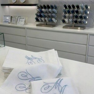 May include: White linen napkins embroidered with a light blue monogram. The napkins are stacked on a white surface. In the background, spools of thread and framed embroidery hoops are displayed on shelves. The store's logo is visible in the background.