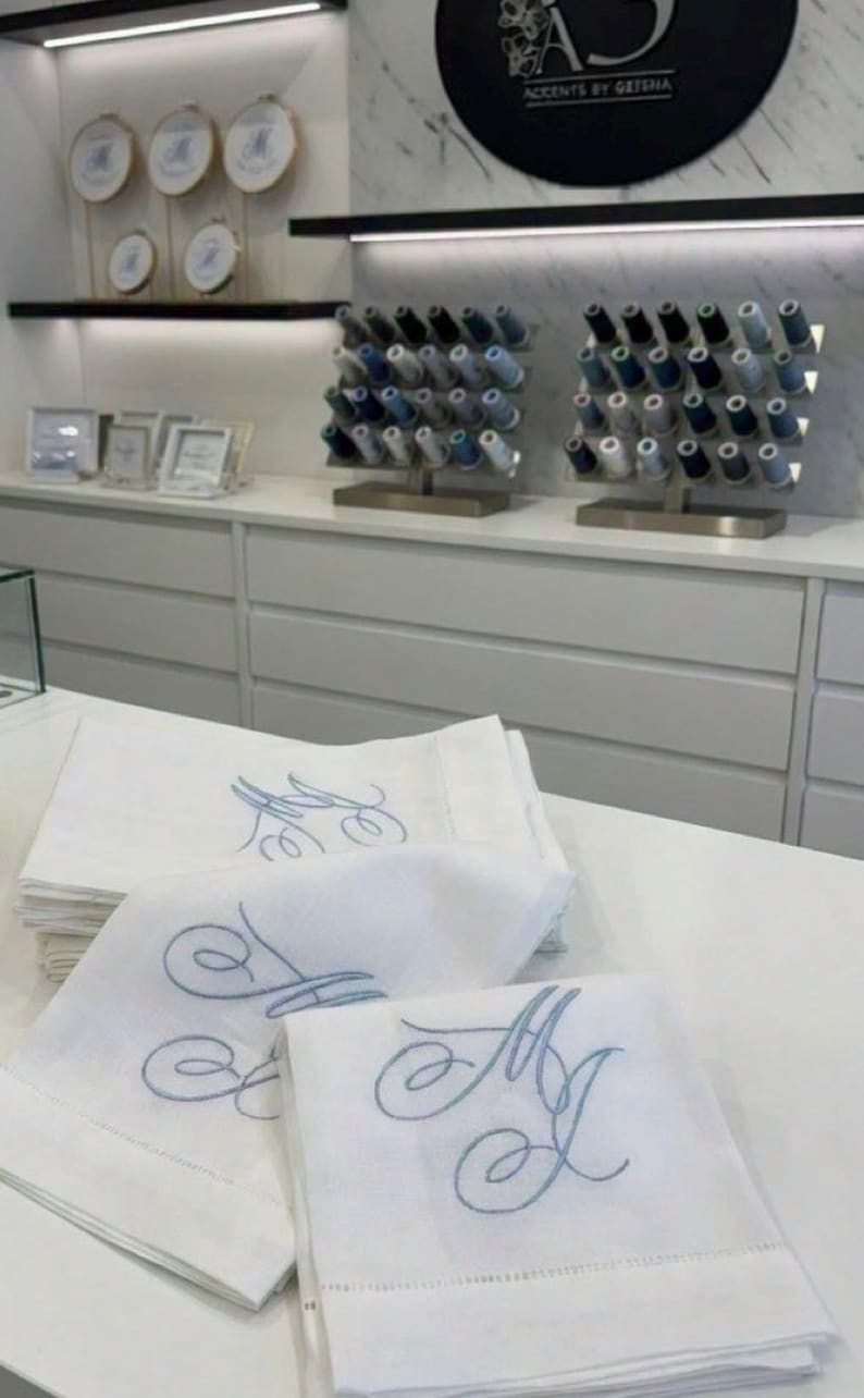 May include: White linen napkins embroidered with a light blue monogram. The napkins are stacked on a white surface. In the background, spools of thread and framed embroidery hoops are displayed on shelves. The store's logo is visible in the background.