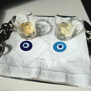 May include: Two clear glass shot glasses filled with a light yellow liquid, sitting on white napkins with blue and white evil eye designs. The glasses are on a silver tray with a silver hook.