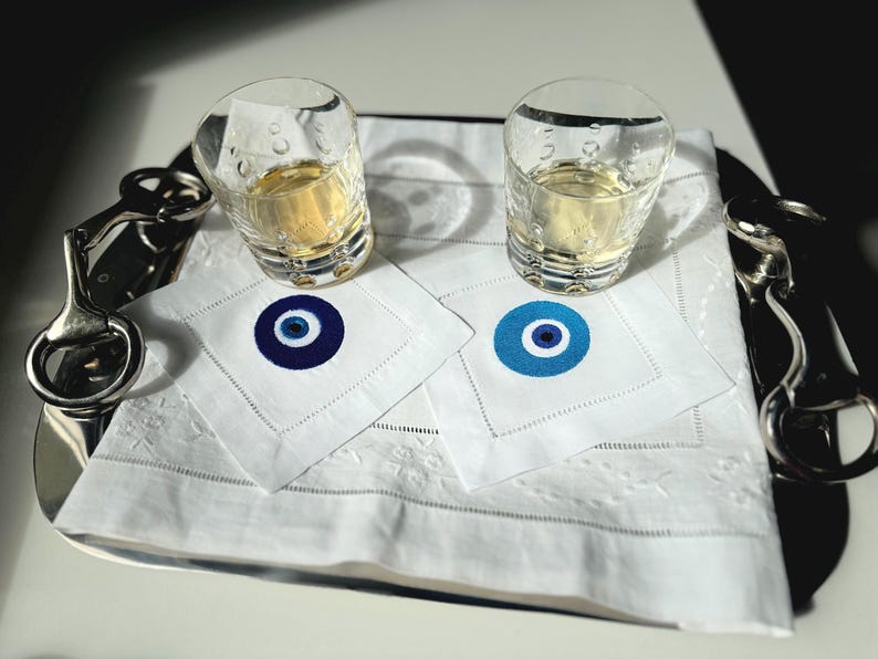 May include: Two clear glass shot glasses filled with a light yellow liquid, sitting on white napkins with blue and white evil eye designs. The glasses are on a silver tray with a silver hook.