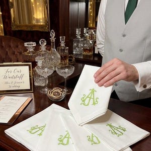 May include: White linen napkins embroidered with a green letter "A". The napkins are displayed on a dark wooden bar, with a person holding one. The scene includes glassware, bottles, and a sign that reads "For Our Waitstaff - Alex & Jessica's Wedding."