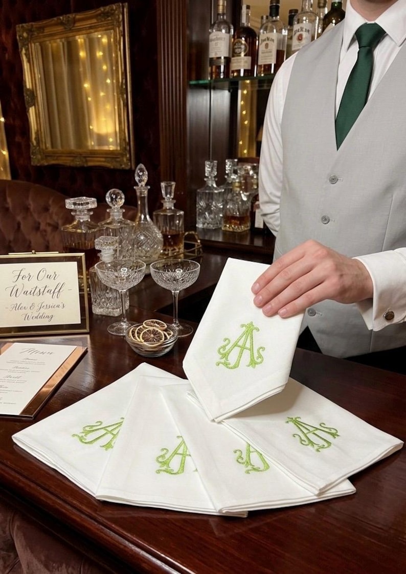 May include: White linen napkins embroidered with a green letter "A". The napkins are displayed on a dark wooden bar, with a person holding one. The scene includes glassware, bottles, and a sign that reads "For Our Waitstaff - Alex & Jessica's Wedding."