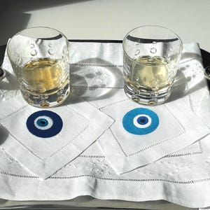 May include: Two clear glass tumblers filled with a light yellow liquid, sitting on white linen napkins with blue and white evil eye designs. The napkins are on a silver tray.