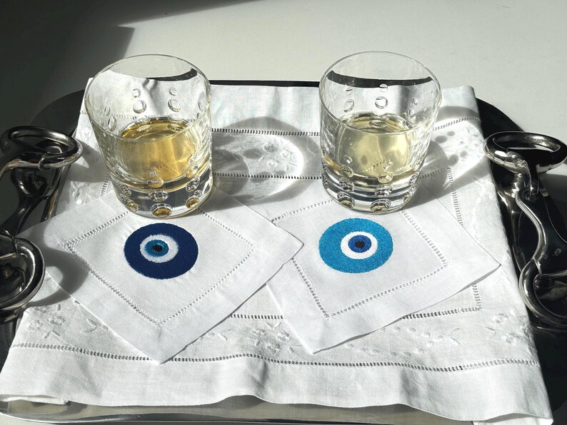 May include: Two clear glass tumblers filled with a light yellow liquid, sitting on white linen napkins with blue and white evil eye designs. The napkins are on a silver tray.