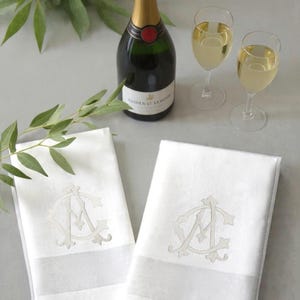 May include: A still-life image featuring two white monogrammed napkins, a champagne bottle, and two glasses of champagne. The napkins have a light gray stripe and a monogram. Green leaves are in the background.