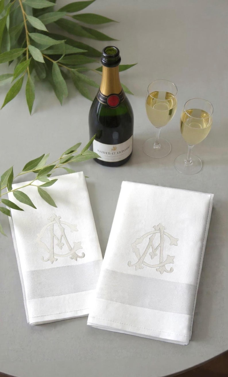 May include: A still-life image featuring two white monogrammed napkins, a champagne bottle, and two glasses of champagne. The napkins have a light gray stripe and a monogram. Green leaves are in the background.
