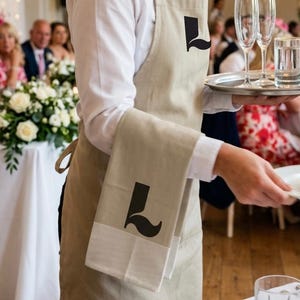 May include: A server wearing a beige apron and matching towel, both with a black "L" design. The server is holding a tray with glasses and a plate, in a well-lit room with a wedding reception in the background.