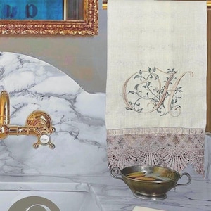 Può includere: Asciugamano in lino color crema con ricamata la lettera "A" e un motivo floreale. L'asciugamano ha un bordo decorativo in pizzo color rosa tenue. Sono visibili anche un rubinetto dorato e una piccola ciotola di metallo.