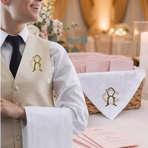 May include: A server in a beige vest and white shirt holds a white towel embroidered with a monogram. A basket with white napkins and a monogram is on a table with pink menus. The setting appears to be a formal event.