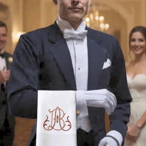 May include: A formal event scene featuring a person in a tuxedo holding a white towel. The towel has a brown monogram design. The person is wearing white gloves and a bow tie. The background is blurred.