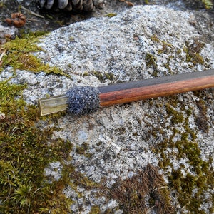 May include: A handcrafted wooden pipe with a dark finish and a rectangular metal end. The pipe features a section covered in small, dark gray stones. The background is a moss-covered rock.