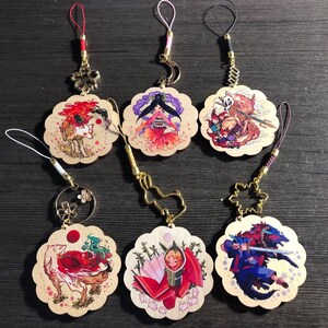 May include: Six wooden charms with colorful illustrations and gold accents. Each charm features a different character or scene, including a fox, a woman in a red dress, a creature with a mask, a woman with a sword, a woman with a fan, and a fox with a red tail. The charms are attached to gold chains with decorative beads.