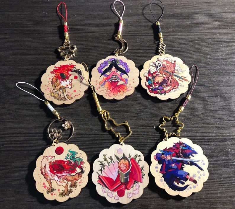 May include: Six wooden charms with colorful illustrations and gold accents. Each charm features a different character or scene, including a fox, a woman in a red dress, a creature with a mask, a woman with a sword, a woman with a fan, and a fox with a red tail. The charms are attached to gold chains with decorative beads.