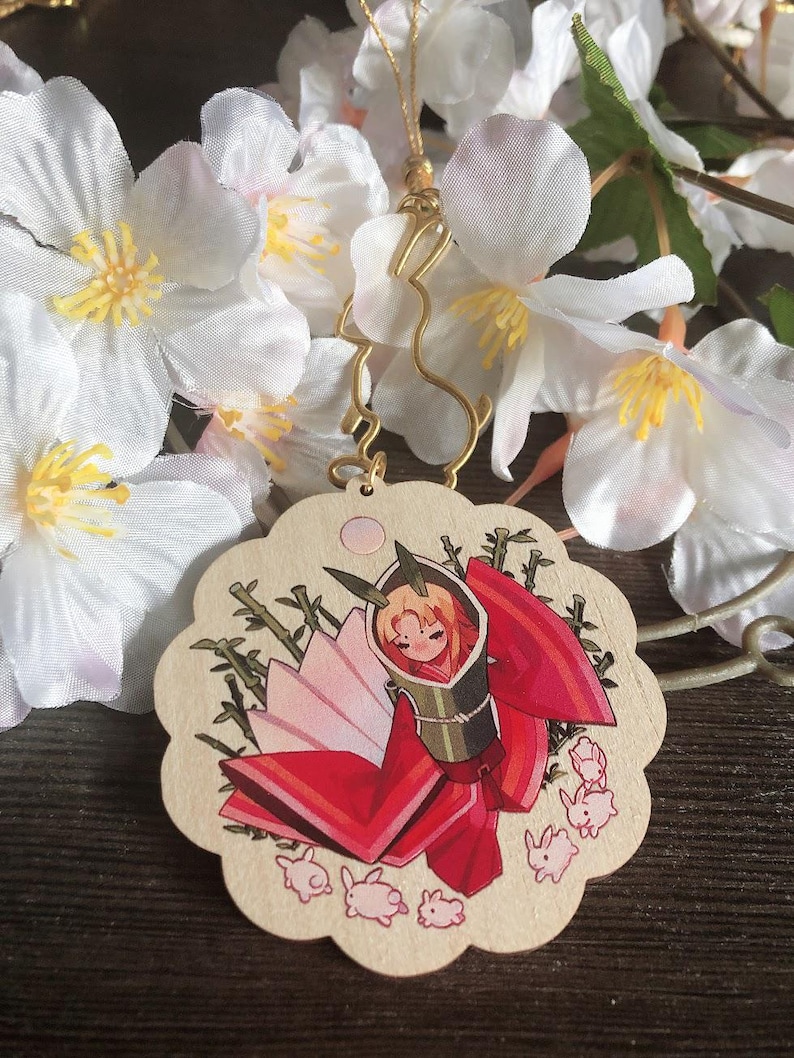 May include: A wooden charm featuring a hand-painted illustration of a woman in a red and white kimono with a bamboo background. The charm is surrounded by small pink rabbits.