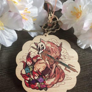 May include: A wooden keychain featuring a red fox with nine tails. The fox is depicted in a Japanese style with a sword and a mask. The keychain is in the shape of a cloud.