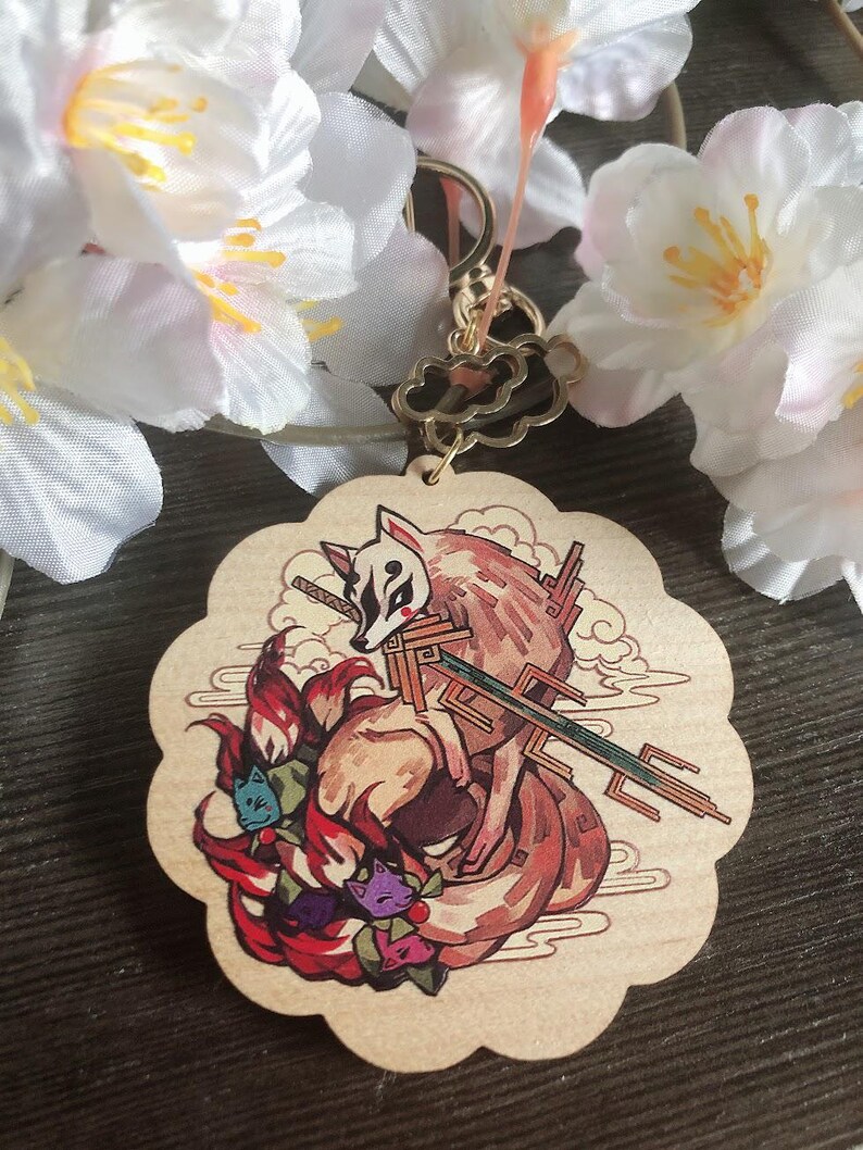 May include: A wooden keychain featuring a red fox with nine tails. The fox is depicted in a Japanese style with a sword and a mask. The keychain is in the shape of a cloud.