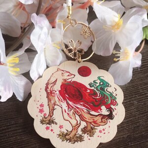 May include: A white wooden charm with a red and green fox design. The fox is surrounded by pink cherry blossoms. The charm is attached to a gold chain with a circle pendant featuring a floral design.