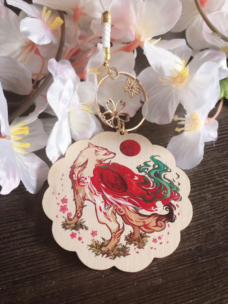 May include: A white wooden charm with a red and green fox design. The fox is surrounded by pink cherry blossoms. The charm is attached to a gold chain with a circle pendant featuring a floral design.