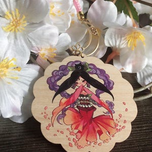 May include: A wooden charm featuring a hand-painted illustration of a woman with long black hair, wearing a red and orange kimono, surrounded by pink flower petals. The charm is attached to a gold chain.