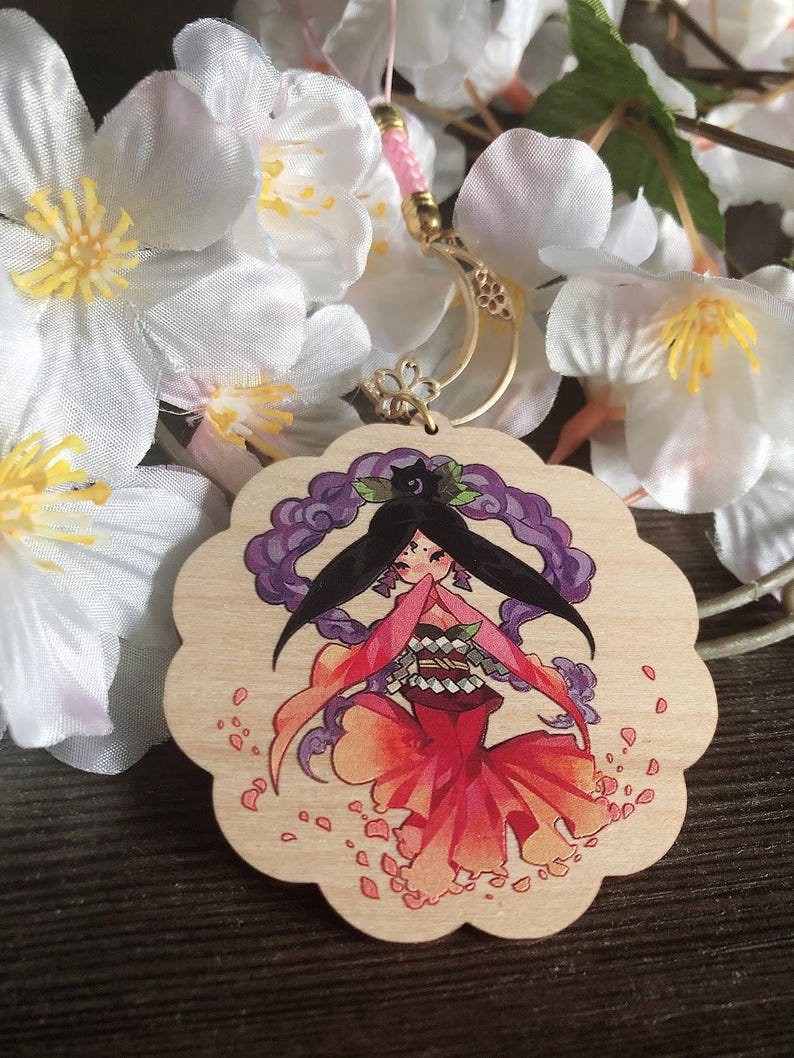 May include: A wooden charm featuring a hand-painted illustration of a woman with long black hair, wearing a red and orange kimono, surrounded by pink flower petals. The charm is attached to a gold chain.