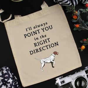 Puede incluir: Una bolsa de tela beige con texto negro que dice "Siempre te señalaré la dirección correcta". Debajo del texto hay una ilustración en blanco y negro de un perro que apunta con el hocico.